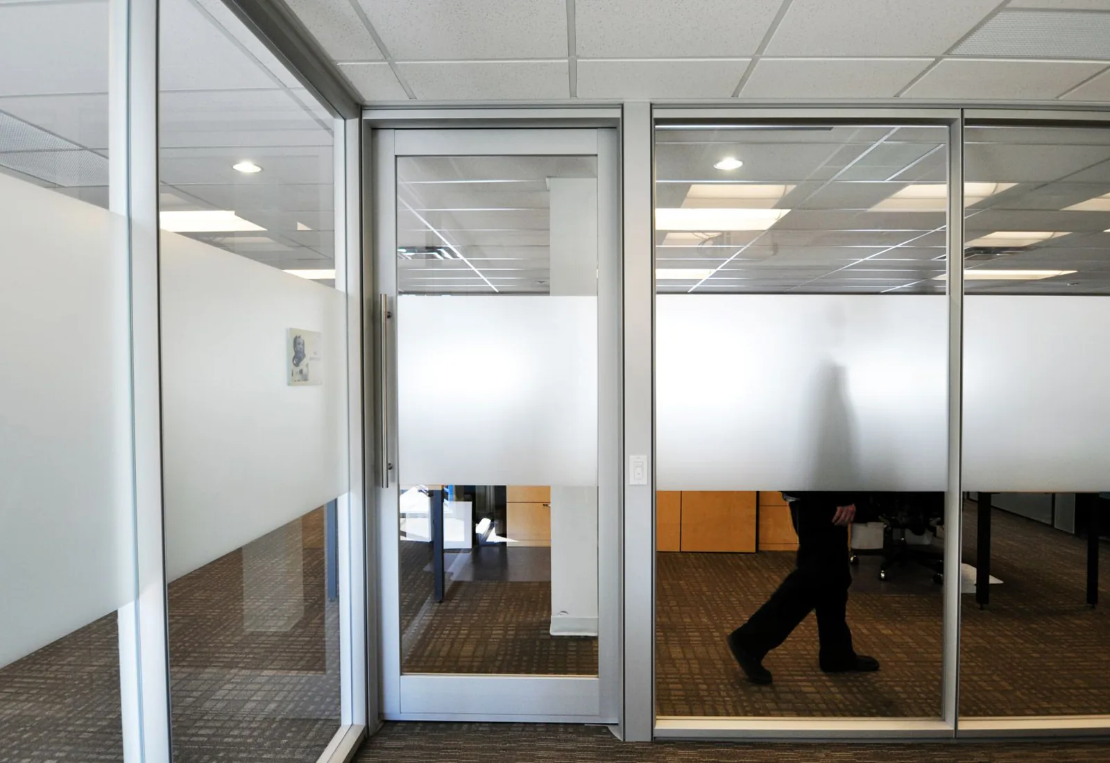Modern office interior with aluminum-framed glass walls, frosted privacy film, and a person walking behind.