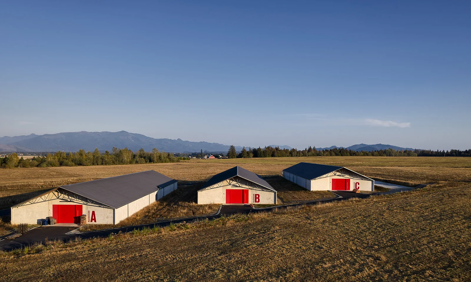 Three modern white rackhouses with bright red doors in a dry field under a blue sky with distant mountains.