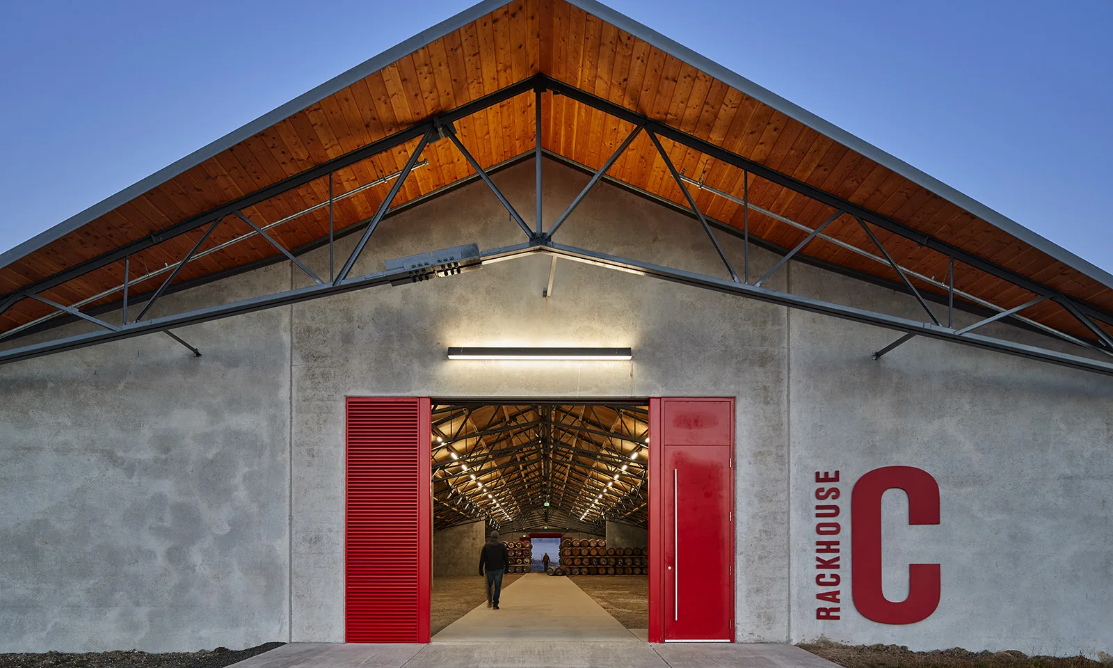 Modern concrete rackhouse with red sliding doors, wood-lined roof, and a view of the barrel storage inside.