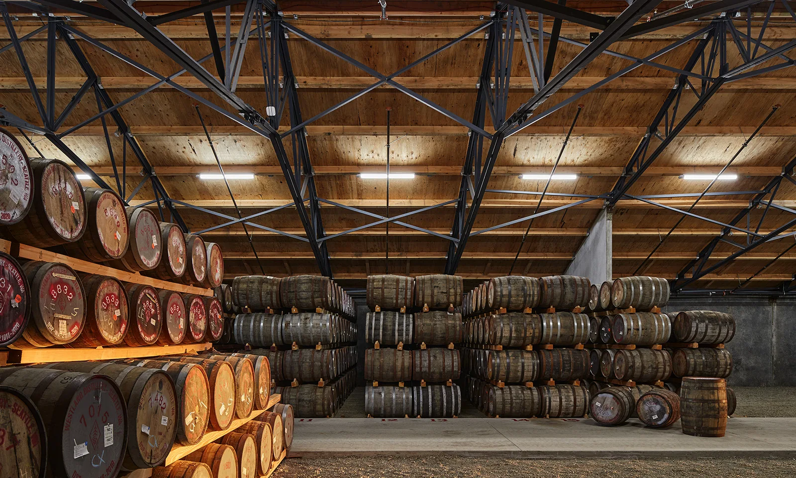 Wooden aging barrels stacked in a distillery warehouse with exposed timber ceilings and steel trusses.