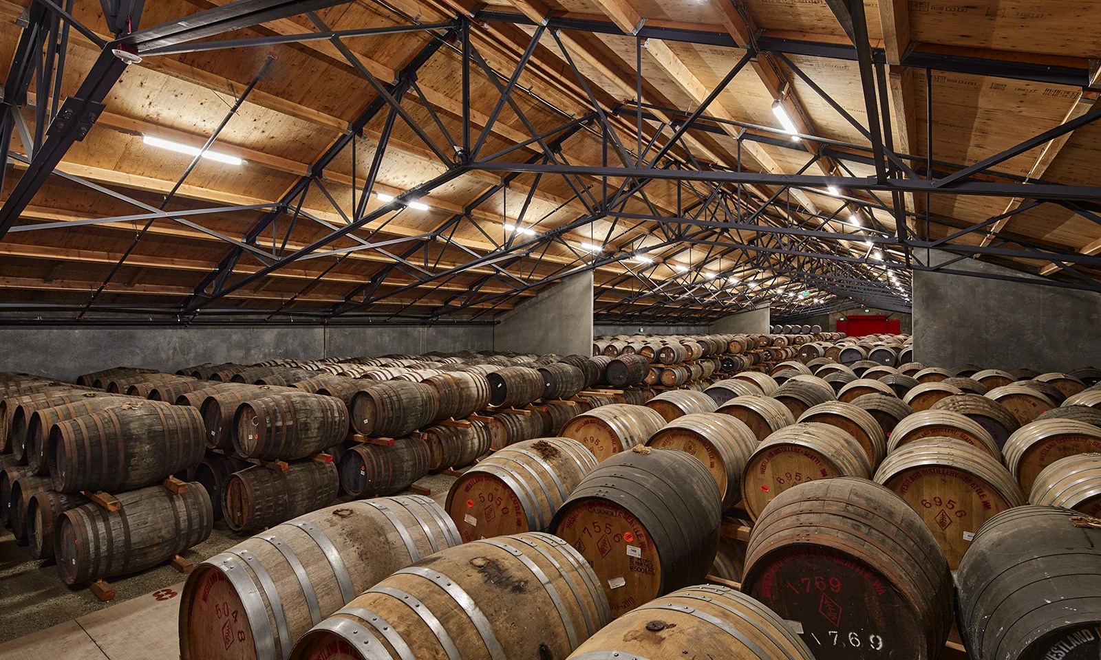 Wooden barrels stacked in a spacious distillery rackhouse with an exposed timber and steel truss ceiling.