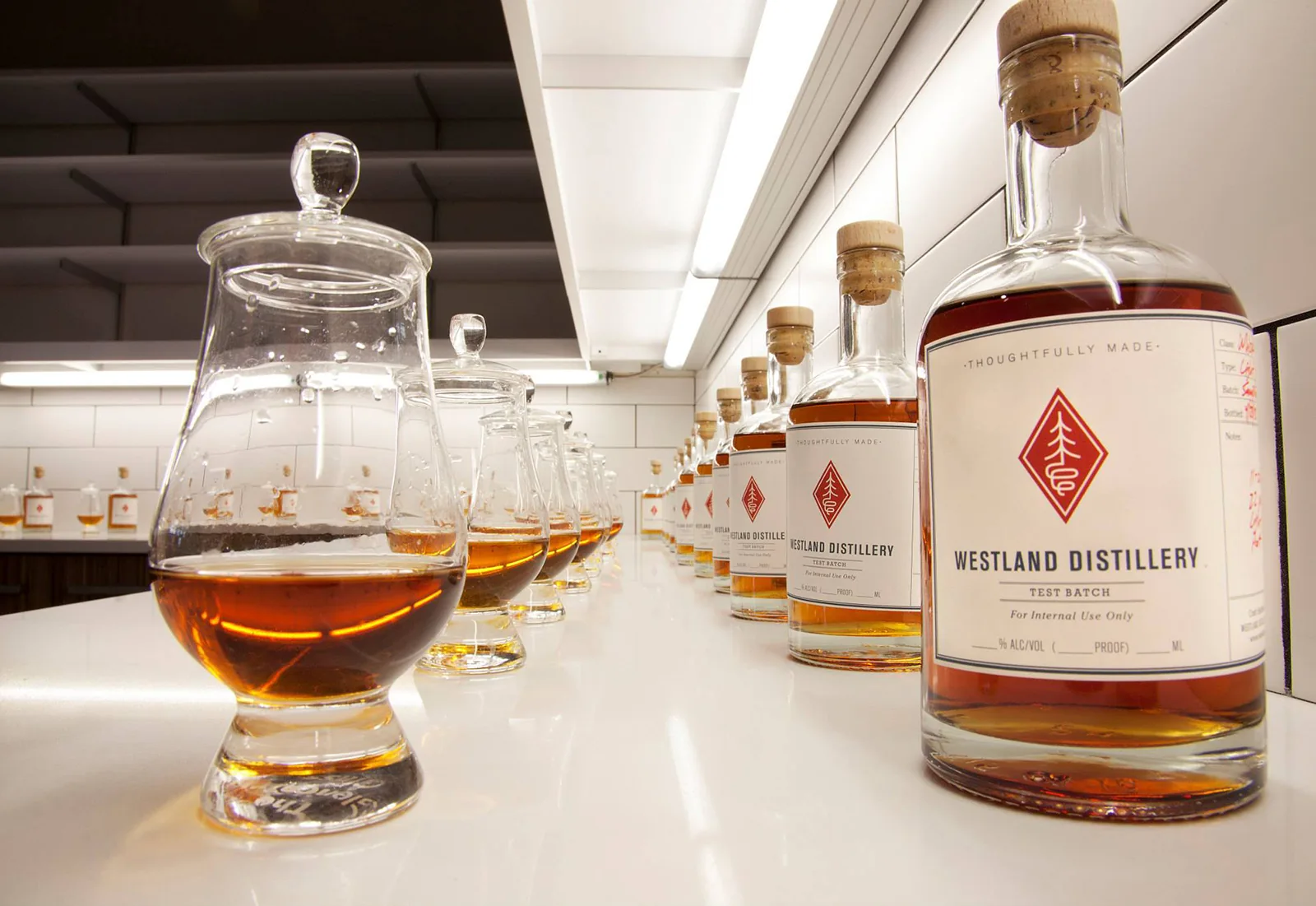 Whiskey tasting glasses and bottles lined up on a clean white countertop in a modern distillery.
