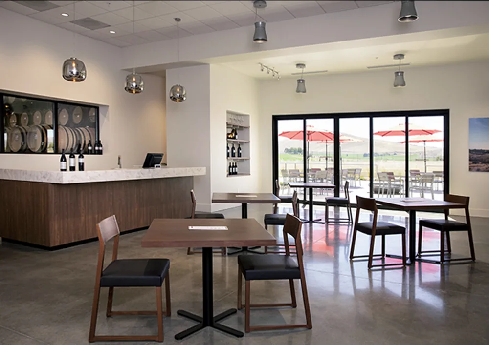 Modern tasting room with polished concrete floors, wooden tables, a marble bar, and glass doors to a sunny patio.