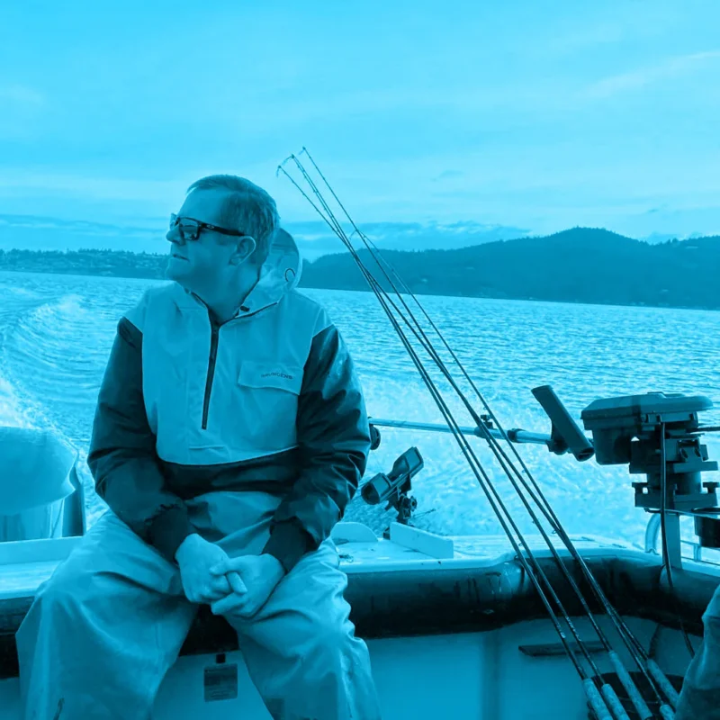 Man in sunglasses sitting on a fishing boat, looking out at the water with hills in the background.