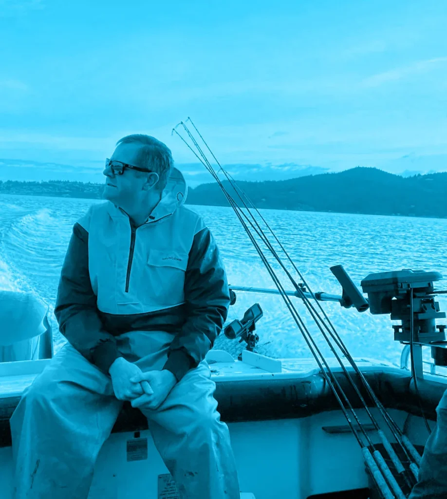 Man in sunglasses sitting on a fishing boat, looking out at the water with hills in the background.