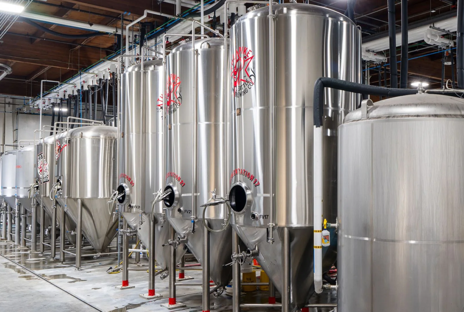 Row of stainless steel fermentation tanks in an industrial brewery with concrete floors and overhead pipes.