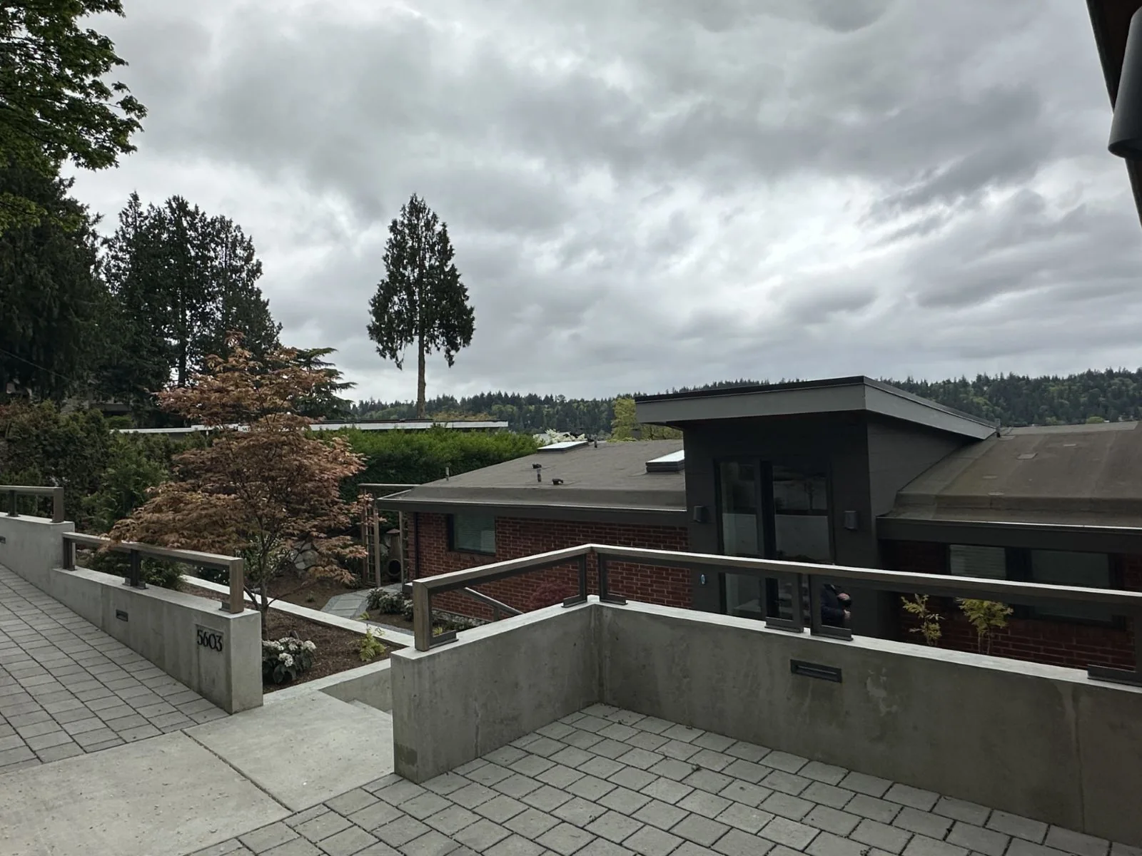 Modern house exterior featuring concrete retaining walls, brick, and dark siding under a cloudy sky.