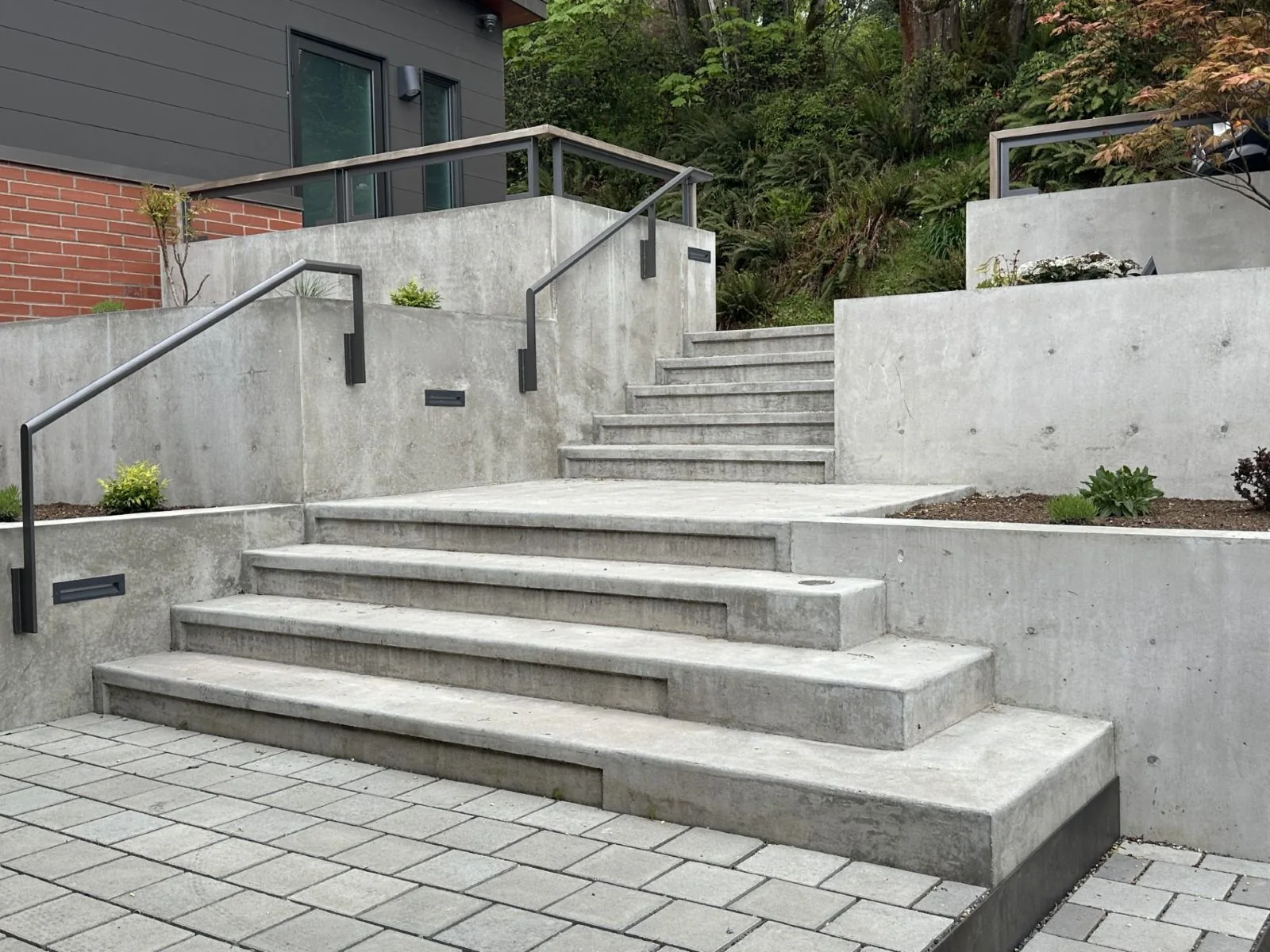 Modern concrete steps and retaining walls with metal handrails leading up a hillside from a paved patio.